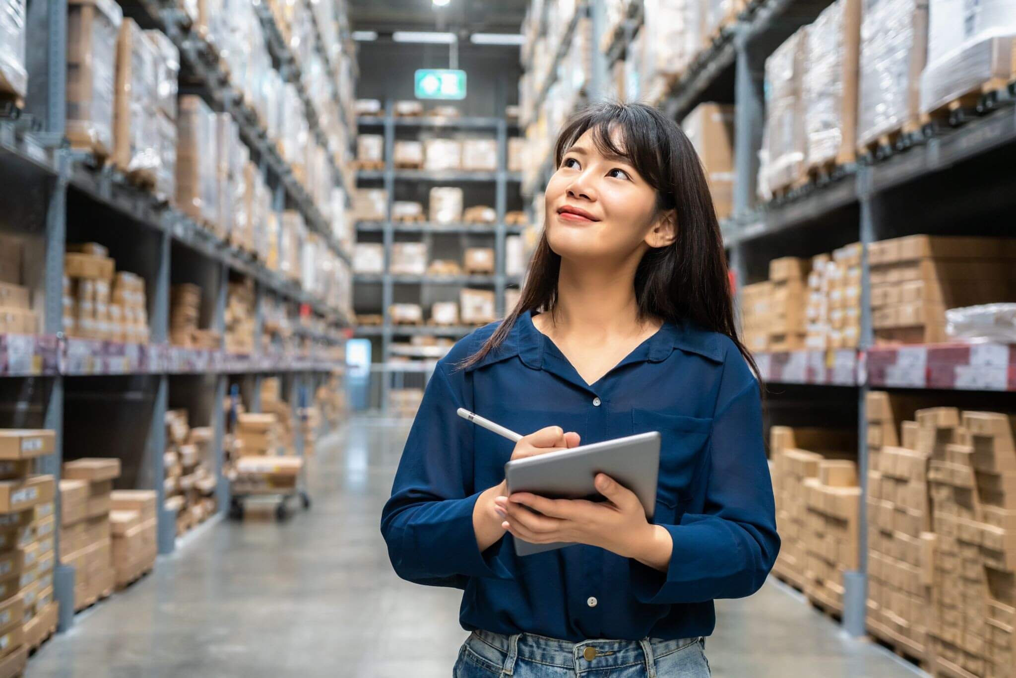 Woman in warehouse holding clipboard looking at cardboard boxes on shelves