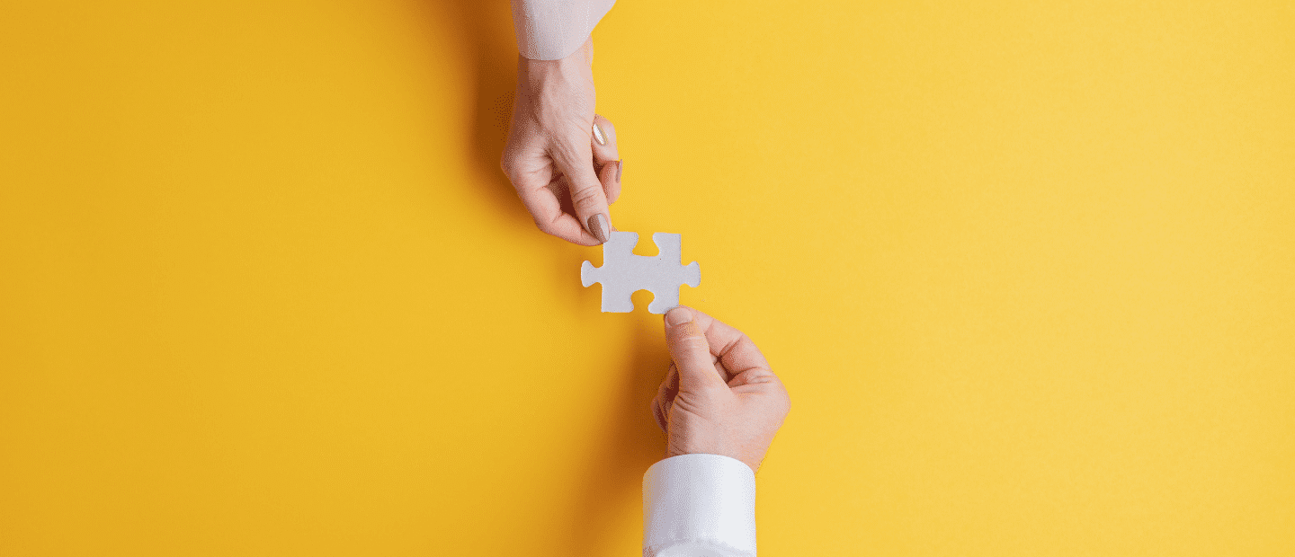 Two hands holding one gray puzzle piece on a yellow background