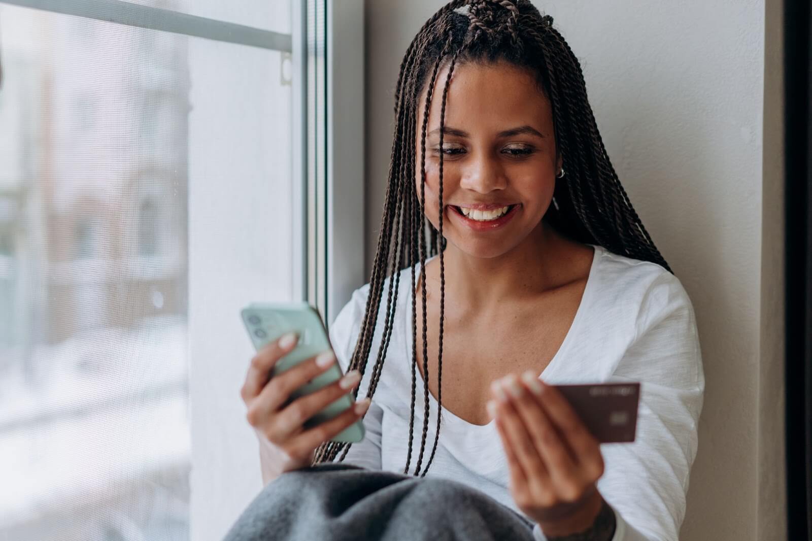Young woman smiling and sitting on a windowsill, looking at her credit card and holding a smartphone in her other hand