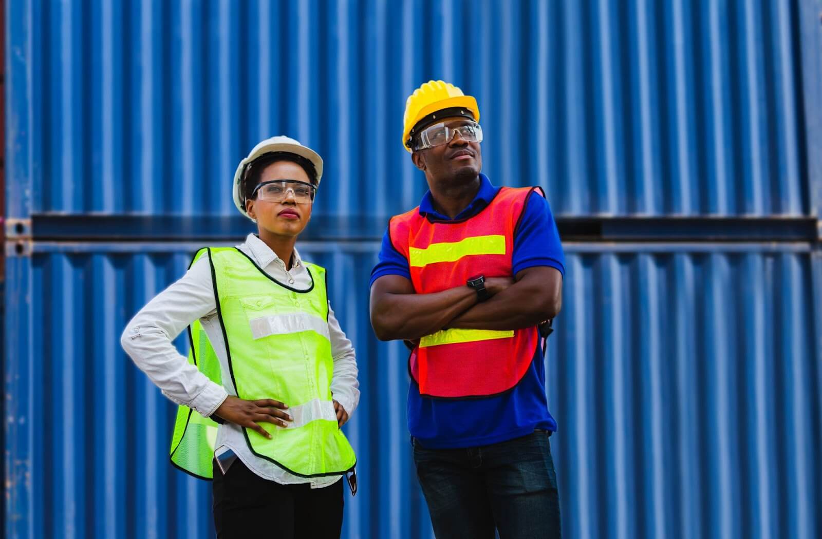 Man and woman wearing reflective vests and hard hats standing in front of a stack of shipping containers