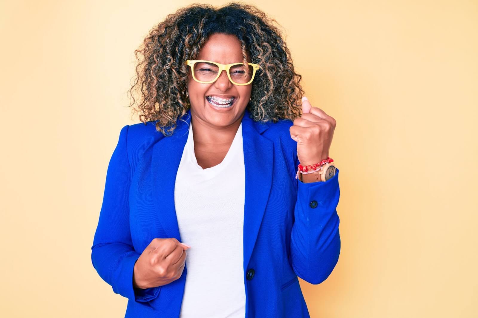 Woman with curly hair wearing bright blue blazer and yellow glasses smiling and pumping her fist in excitement
